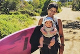 Mez Maconochie carries a baby in a harness while holding a surfboard.