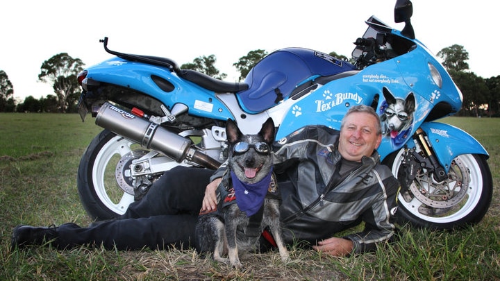 A smiling, grey-haired man and his cattle dog, both dressed in riding gear, posing in front of a motorcycle.