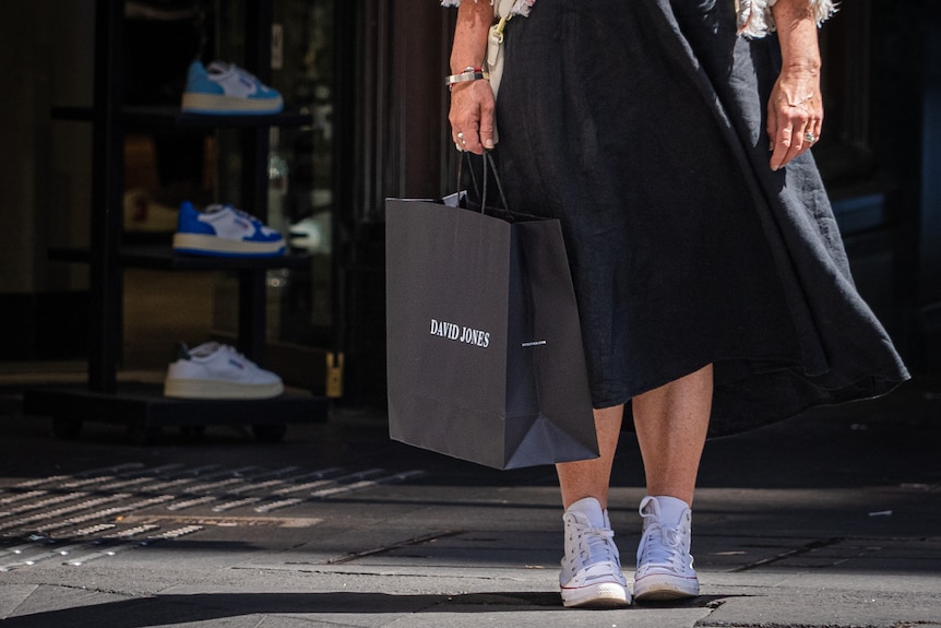 A woman from her waist down, wearing a long black dress and white sneakers holding a black shopping bag that says 'David Jones'