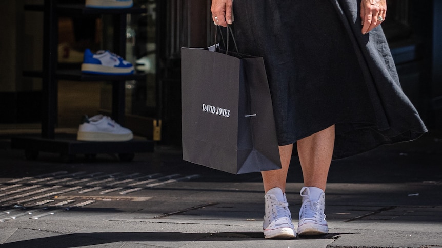 A woman from her waist down, wearing a long black dress and white sneakers holding a black shopping bag that says 'David Jones'