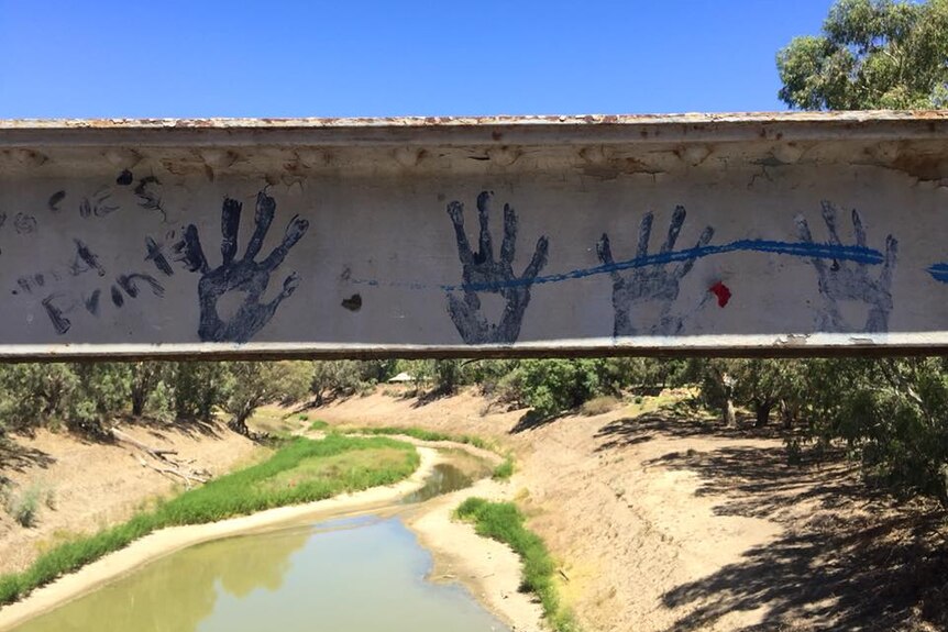 The handrail of the bridge has blue handprints on it from the community and behind it lays a green murky muddle in the river