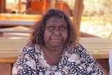 Bessie Nipper is wearing a floral shirt and looking at the camera. Behind her is red dirt and picnic tables.