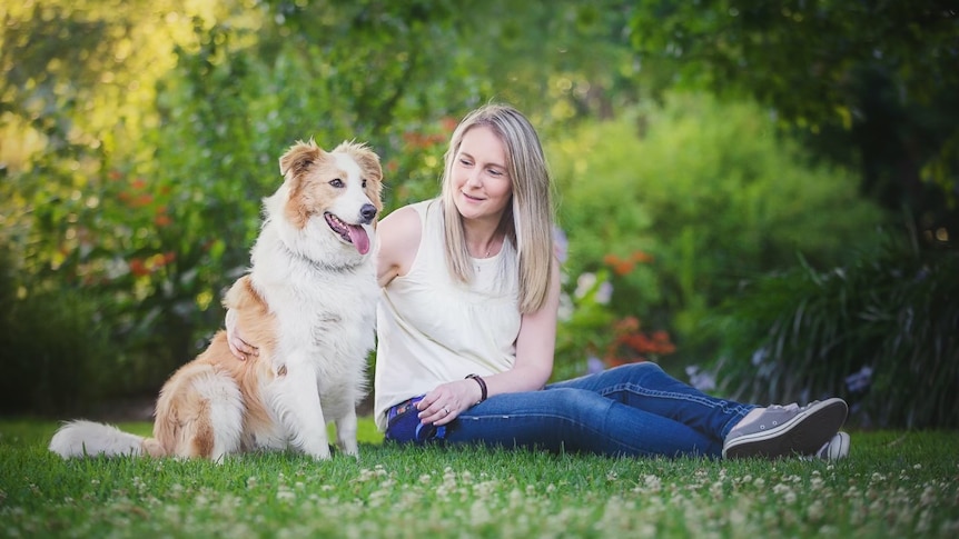 A woman with long blond hair wears a singlet and jeans and is sitting in a park with her arm around a border collie.