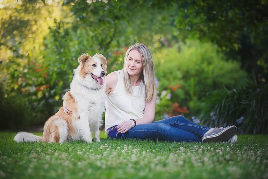Kate has long blond hair and is wearing a singlet and jeans and is sitting in a park with her arm around a border collie