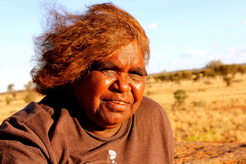 Portrait of Aboriginal elder Monica Robinson. Sunny day and dry bush landscape in the background.