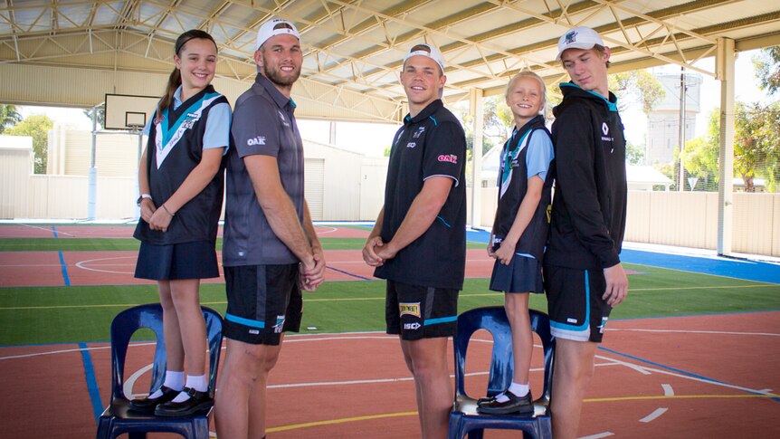 Students Nikki and Paityn stand on chairs next to Port Adelaide players Jackson Trengrove, Dan Hewson and Todd Marshall.