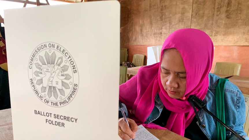 A bright wood-panelled schoolroom is turned into a polling station, with a voter in a bright pink hijab writing on polling paper