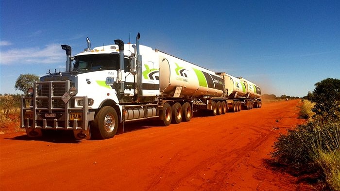 A large white truck with three trailers on a red dirt road under clear blue skies.