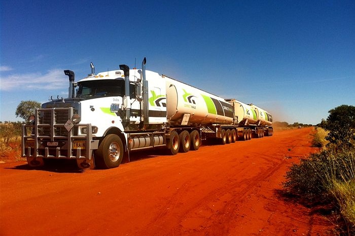 A large white truck with three trailers on a red dirt road under clear blue skies