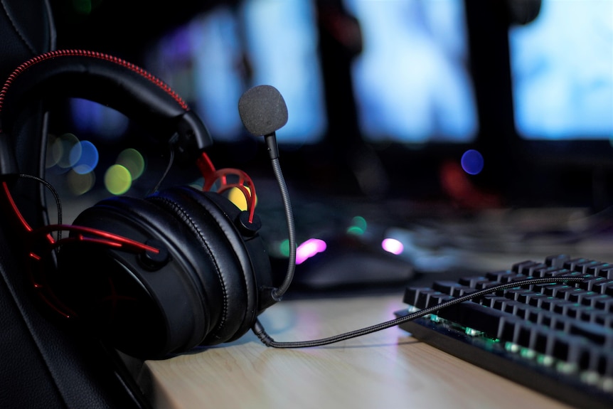 Headphones sit on a computer desk. A neon keyboard and computer sit behind it.