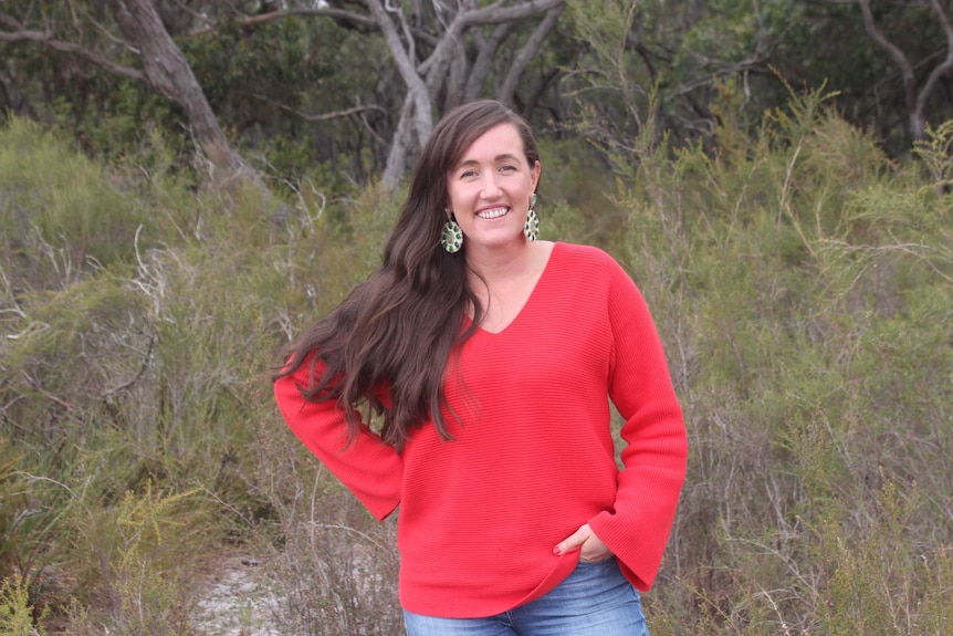 A woman with long brown hair, wearing a red top, standing in front of scrubby trees.