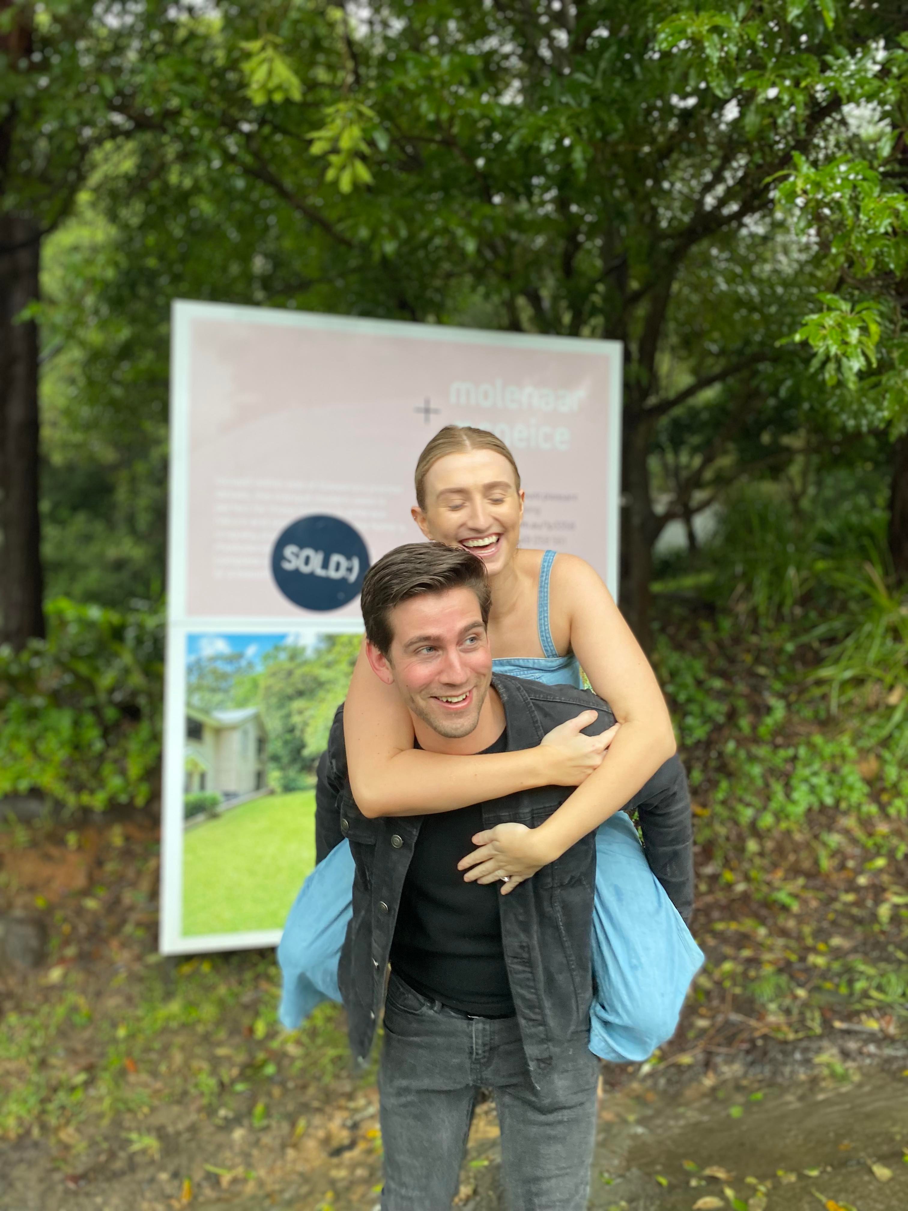 Tyler gives Jess a piggy back while standing in front of a real estate sign.