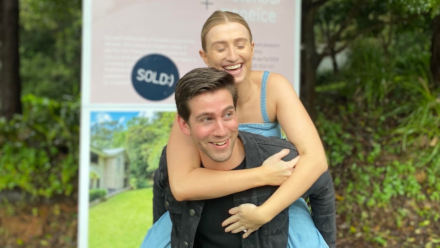 Tyler gives Jess a piggy back while standing in front of a real estate sign, which says sold.