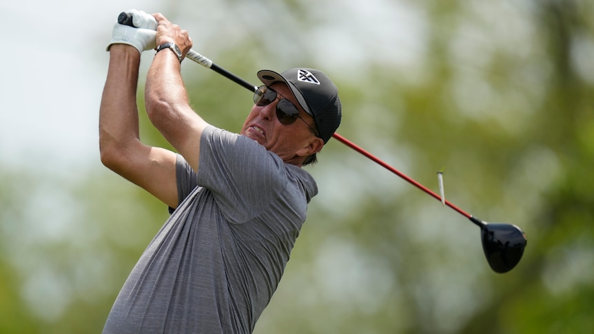 A middle-aged white man in a grey polo shirt and dark cap swings a golf club.