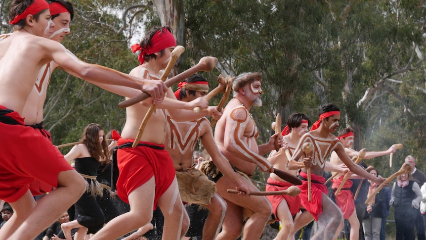 Dancers in traditional costume and body paint dance, carrying sticks on sand at a nature reserve as people look on.