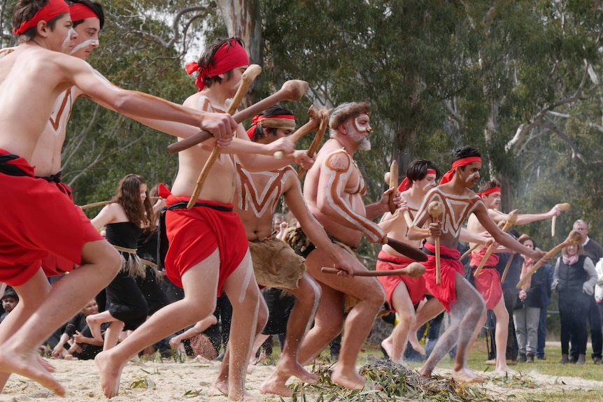 Dancers in traditional costume and body paint dance, carrying sticks on sand at a nature reserve as people look on.