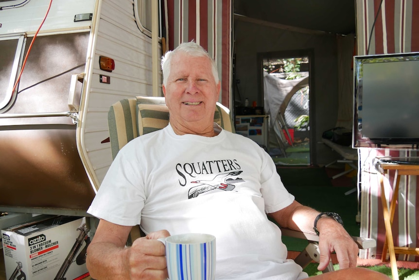 A smiling middle-aged man drinks a cup of tea while sitting in a deckchair outside a caravan