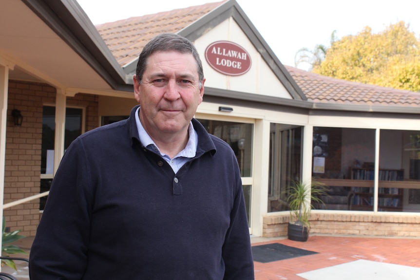 A man in a navy jumper stands in front of a aged care home.