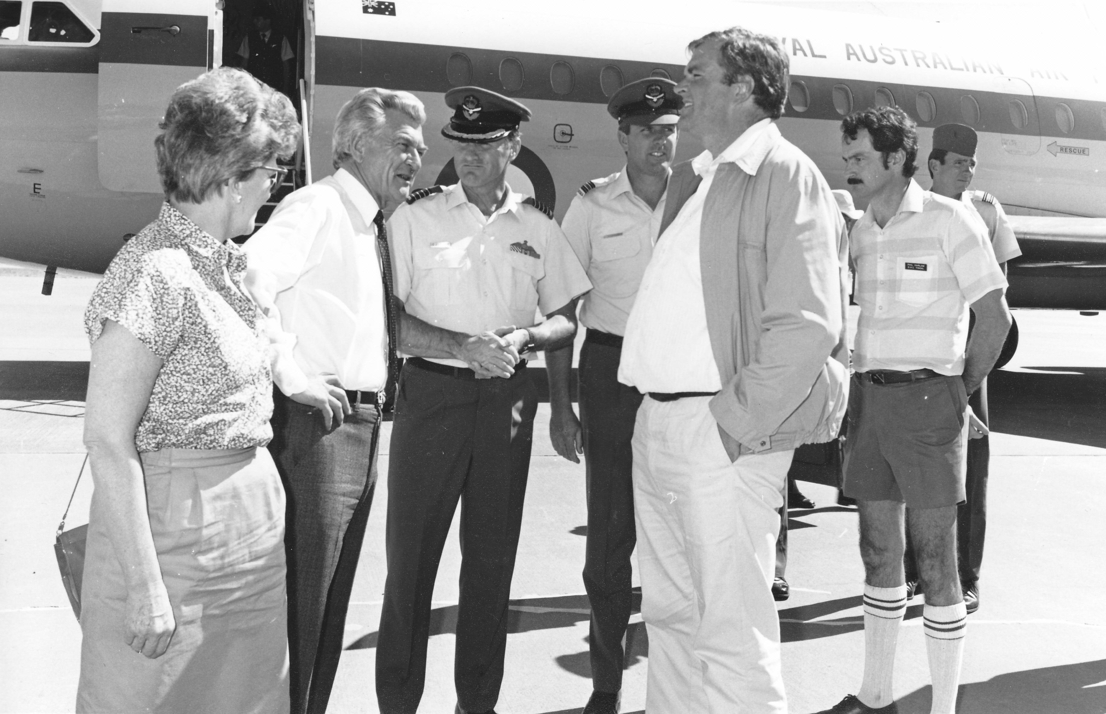 seven people stand as if talking to one another in front of a plane, in black and white
