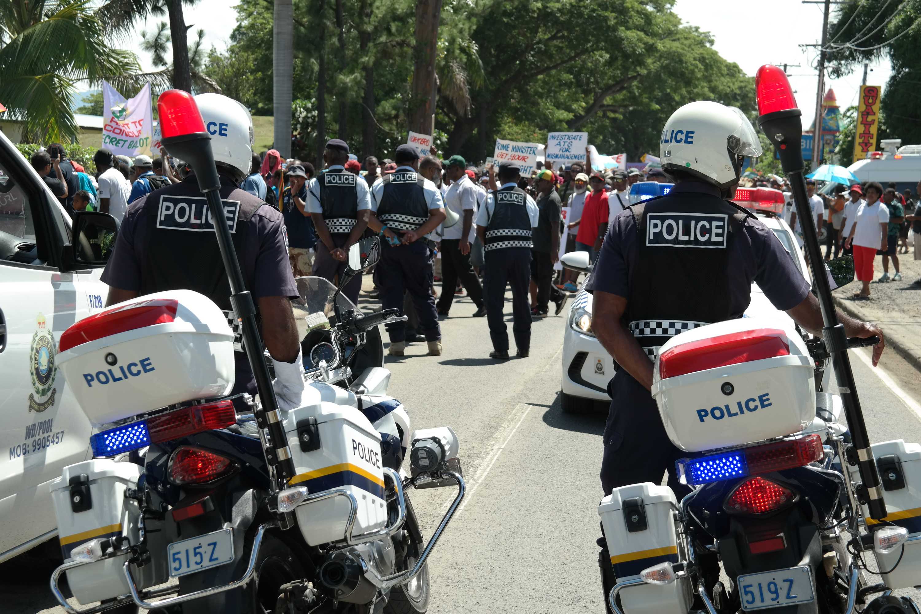 Police cars line a street in Nadi, Fiji, as protestors walk past.