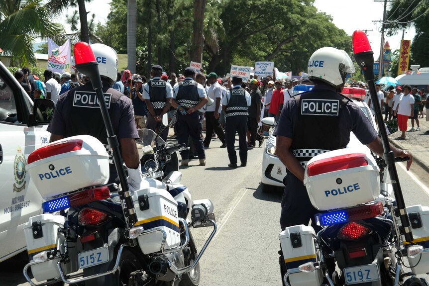 Police cars line a street in Nadi, Fiji, as protestors walk past.