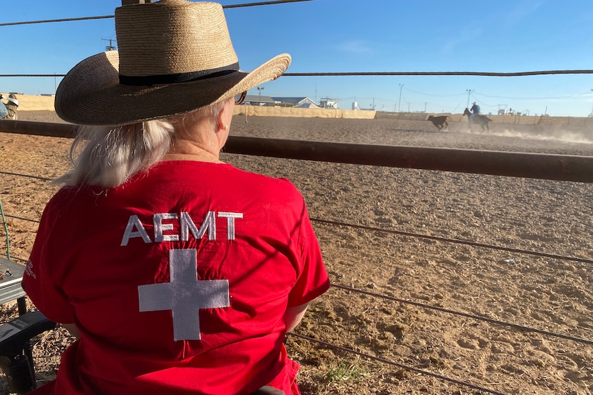 We can see the back of a woman wearing red scrubs and a hat, looking out over a dusty brown campdrafting arena.
