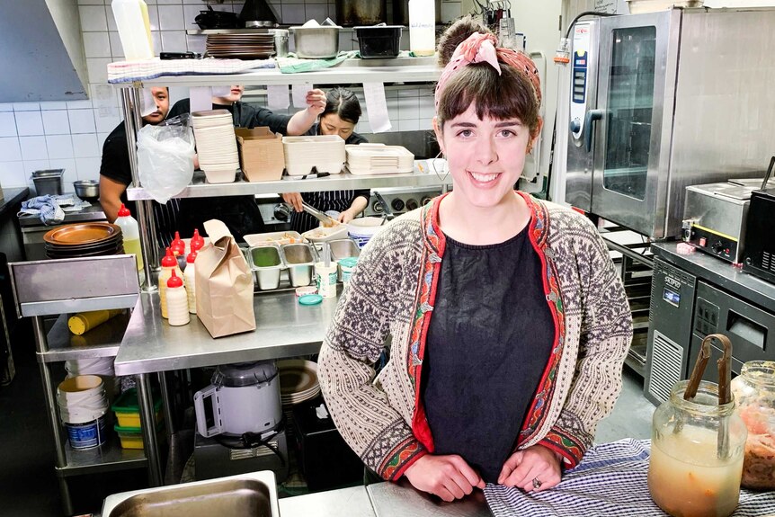 A woman stands in a kitchen where chefs are preparing good.