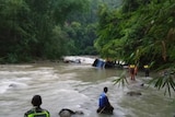 Looking across a fast-flowing river, rescue workers approach a submerged bus as they are tethered with rope.