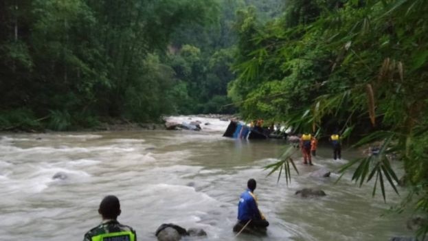 Looking across a fast-flowing river, rescue workers approach a submerged bus as they are tethered with rope.