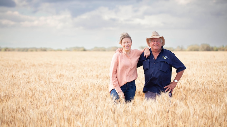 A woman stands in a wheat crop paddock and is leaning on a man wearing a hat