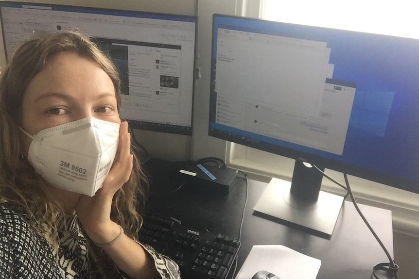 A woman wearing a respirator mask sits in front of two computer monitors