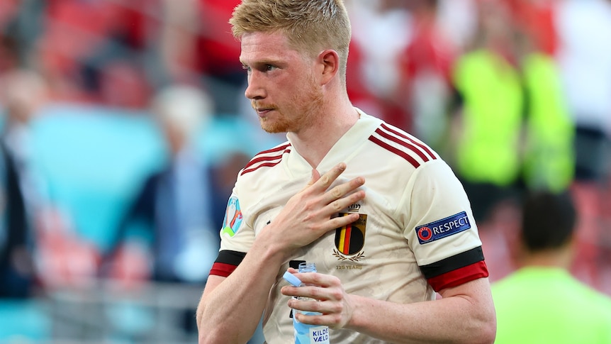 Kevin De Bruyne holds one hand to his chest and a water of bottle in the other