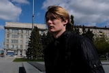 A man stands outside in a public square in Russia, there's a Soviet-style grey block building behind him.