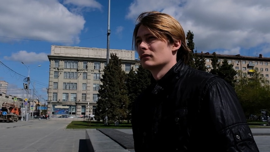 A man stands outside in a public square in Russia, there's a Soviet-style grey block building behind him.