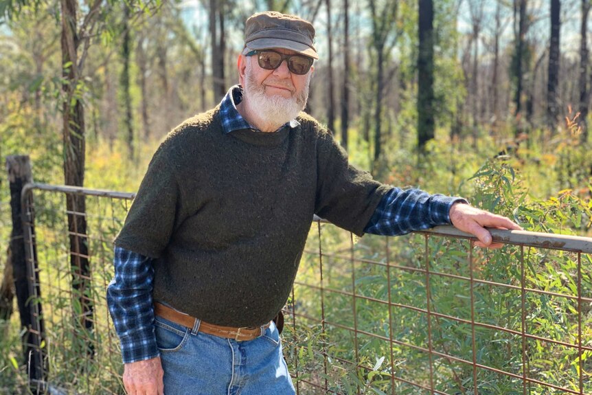 A man with a beard and sunglasses leaning against a fence.