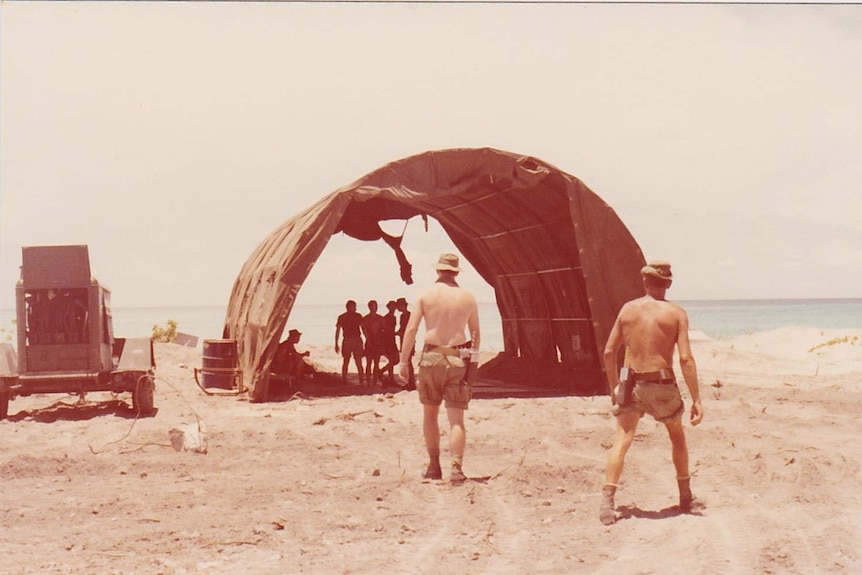 Shirtless soldiers walk towards a shelter where a group of soldiers have gathered out of the sun.