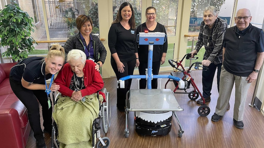 A blue and grey robot surrounded by staff and elderly people