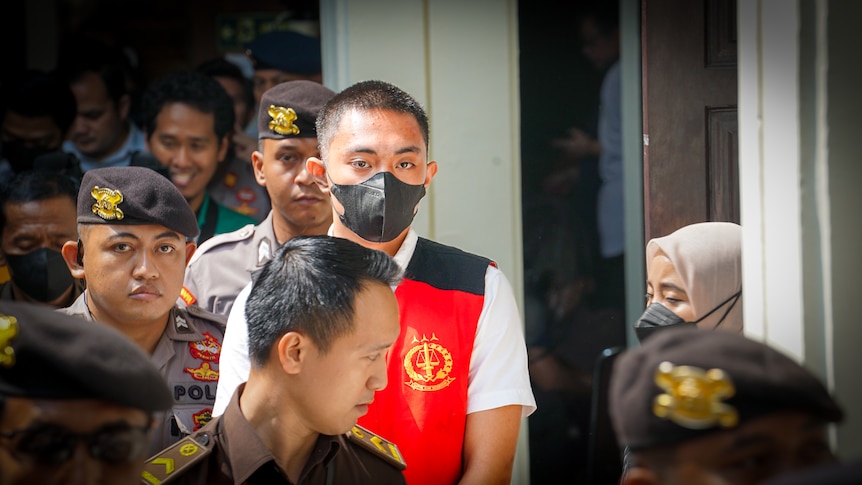 A young man in a black face mask walks while surrounded by Indonesian police