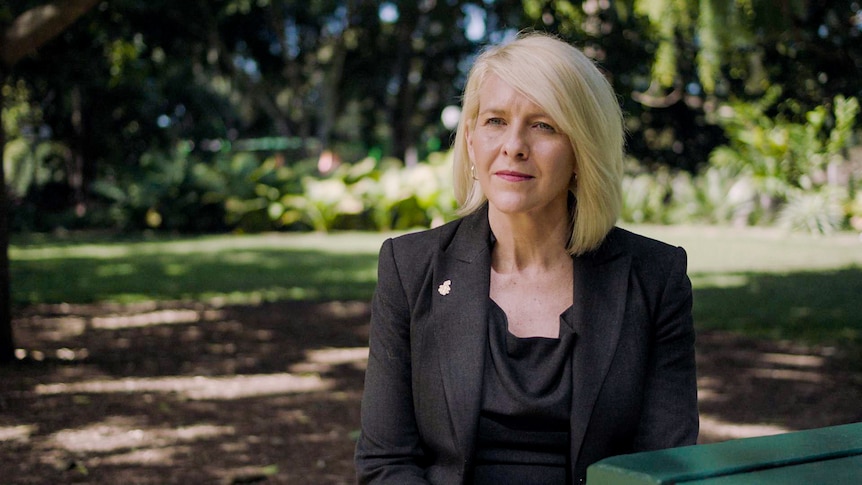 A woman in business attire sits outside, a garden behind her.