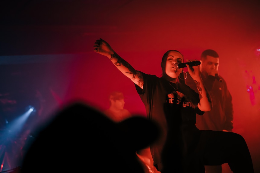 Woman with tattoos wears black Adidas shirt and sings into microphone on a red stage. Man with short hair in jacket sings behind