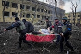 Five men using a stretcher to carry a pregnant woman through rubble outside of a damaged hospital building.