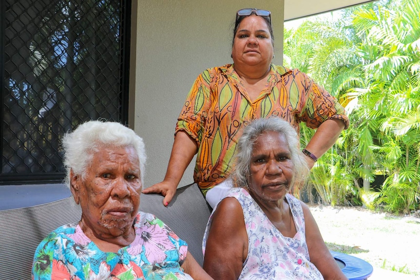 Two elderly indigenous women sit at a table with a younger indigenous woman from their family standing behind them.