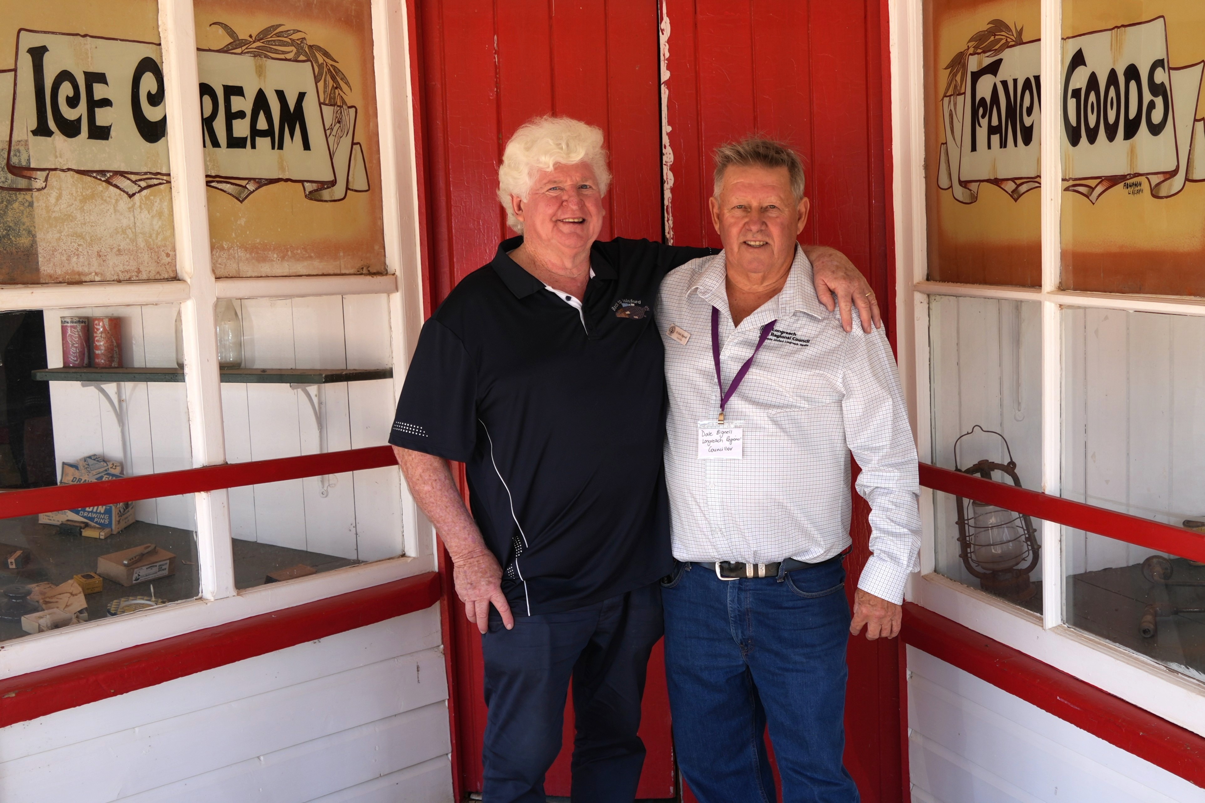 A man with white hair and an Isisford polo shirt poses with a man with a Longreach council shirt in front of a vintage red door.