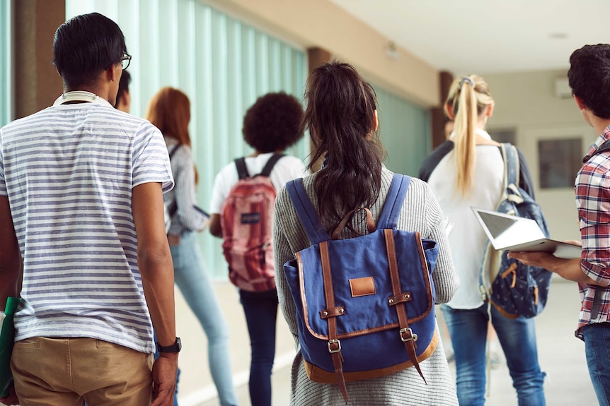 A rear view of a group of young people wearing casual clothes, with backpacks, school books or laptops, in a hallway.