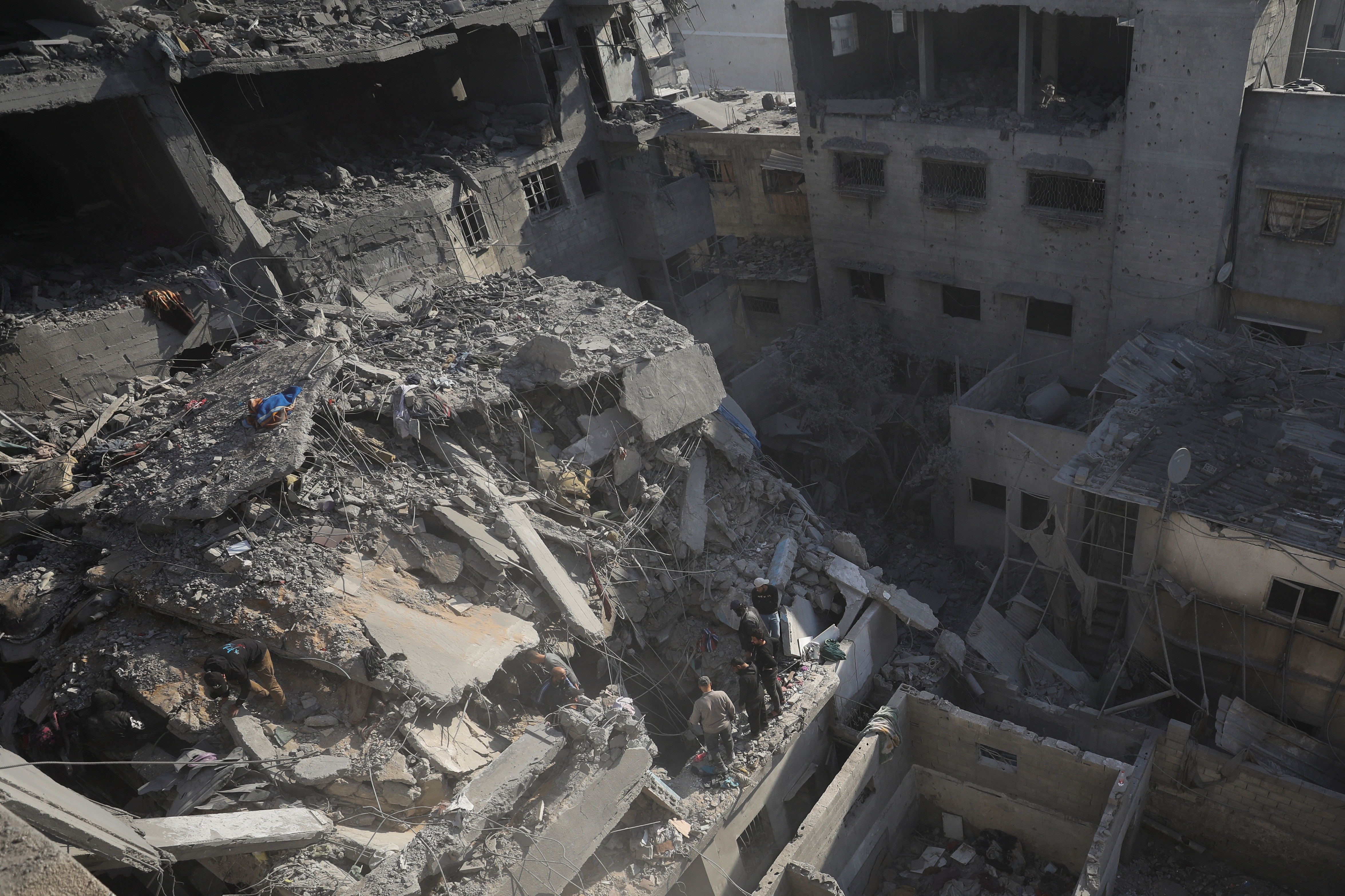 An aerial view of several apartment blocks in ruins with cascading grey rubble, as three people stand at the edge and look