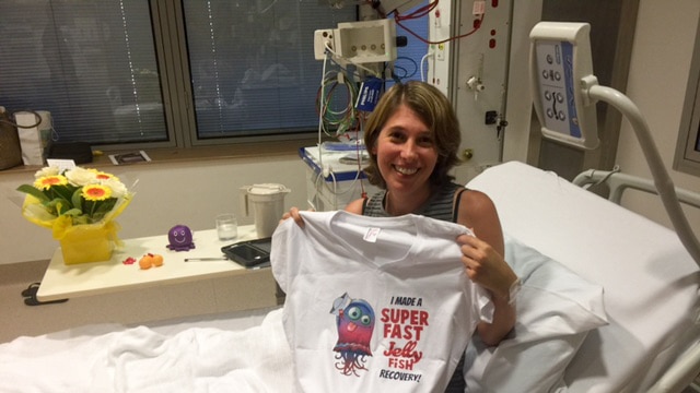 A woman sits on a hospital bed holding a t-shirt emblazoned with the slogan 'I made a super fast jellyfish recovery'.