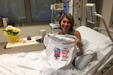 A woman sits on a hospital bed holding a t-shirt emblazoned with the slogan 'I made a super fast jellyfish recovery'.