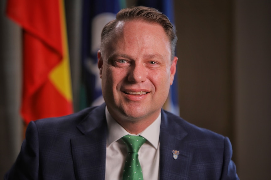 A man wearing a dark suit sits smiling in front of some flags.