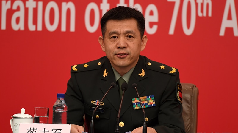 A Chinese man in uniform sitting in front of a red backdrop speaking into a mike.