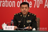 A Chinese man in uniform sitting in front of a red backdrop speaking into a mike.